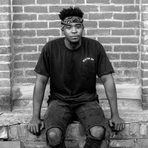 An African American man, sitting on an old brick wall wearing a black tshirt, bandana around his forehead, with tall, thick curly hair. person: austin parker