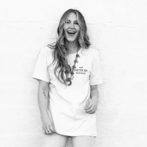 A happy Caucasian female with long medium-toned curly hair, wearing a white Scatter Joy Project tshirt—leaning against a white brick wall. person: Bay Moraghan