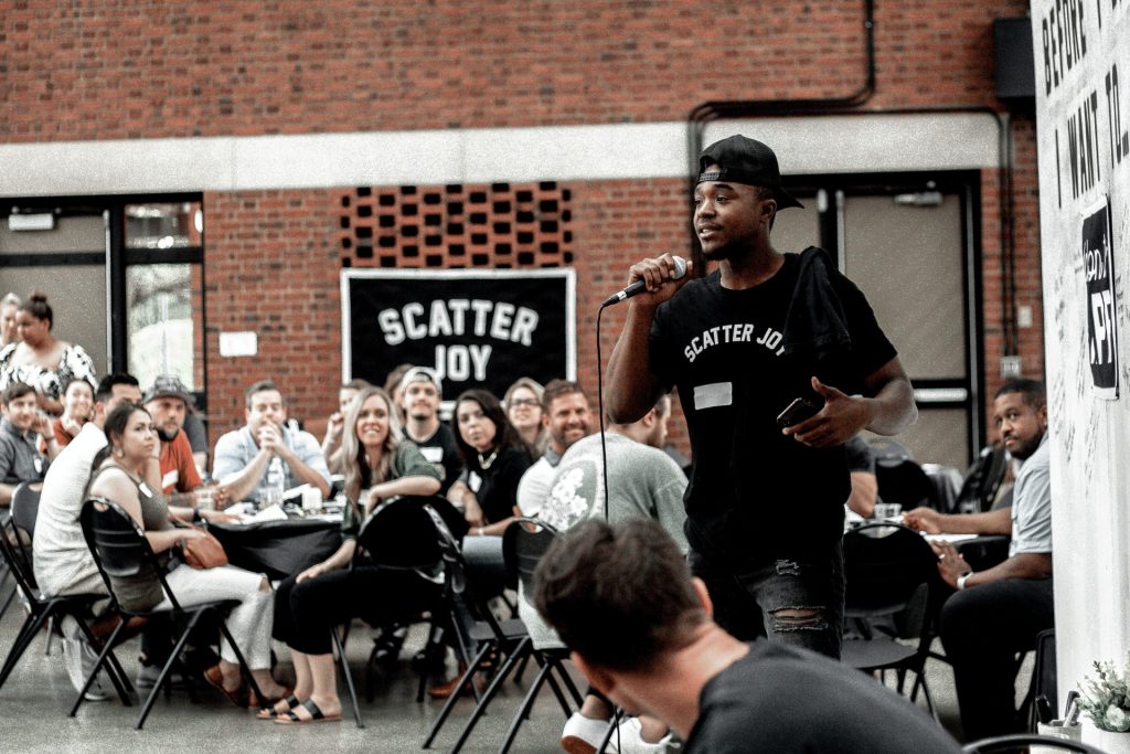 An African American man in Scatter Joy Project attire, giving a live speaking event to a group of people.