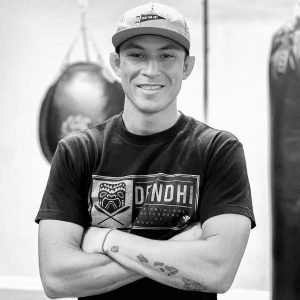 Young Pacific Islander male with Scatter Joy Project baseball hat and black tshirt, standing in a gym with punching bags and equipment, smiling. Person: martin day