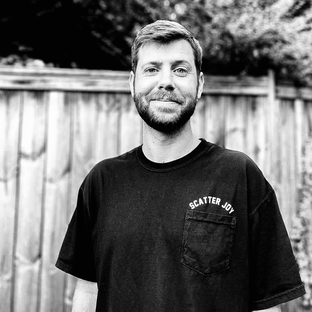 A Caucasian male with short hair and beard, wearing a black Scatter Joy Project tshirt—outside in front of a fence. Person: tanner olsen