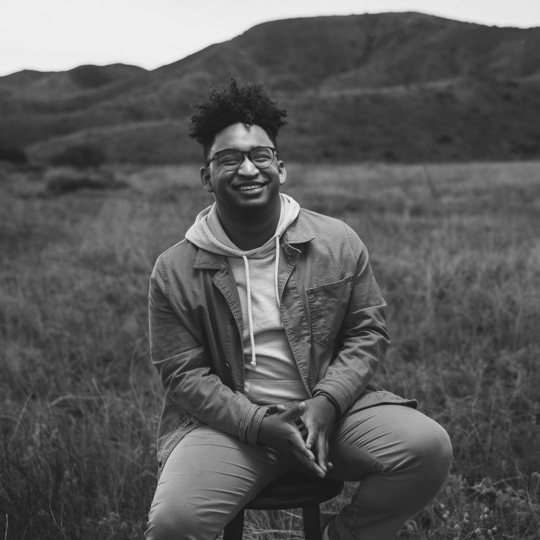 A black and white image of a young African American man with thick, curly hair outside in a field, smiling. Person: Donovan Beck