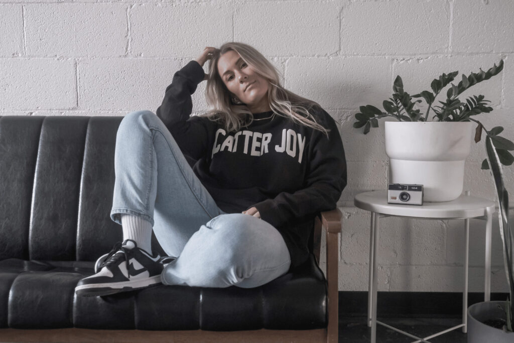 A young Caucasian woman with long, blonde hair wearing jeans and a black Scatter Joy Project sweatshirt—sitting on a black leather sofa.